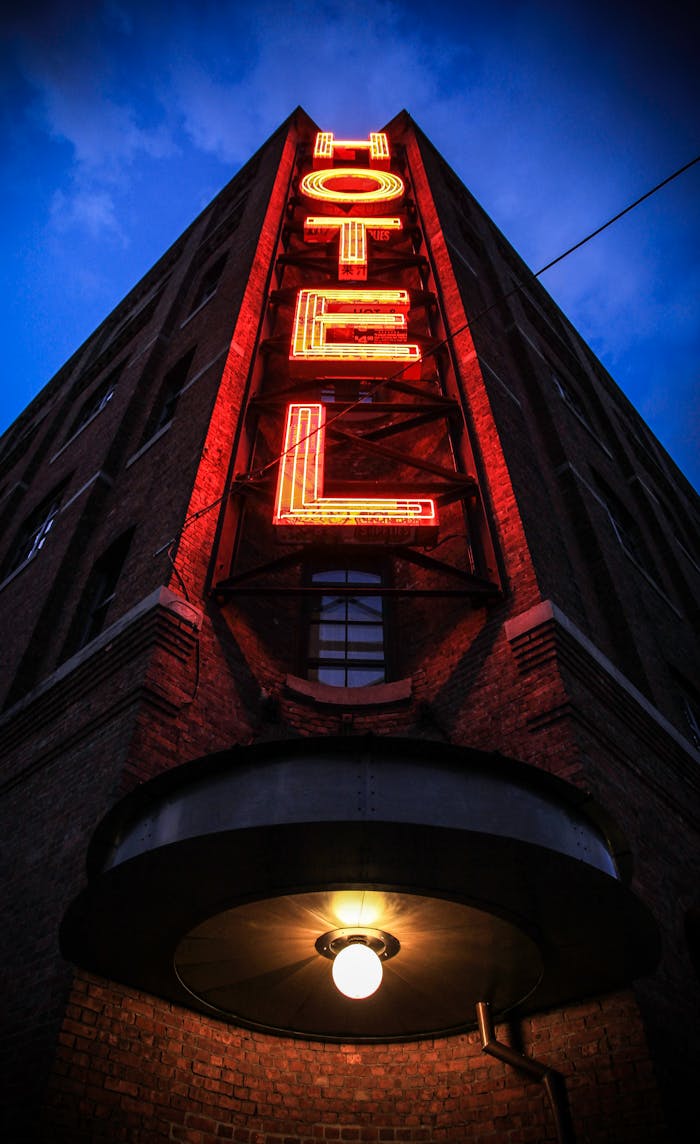 contact-bg Eye-catching low angle shot of a neon hotel sign illuminating a brick building facade at dusk.