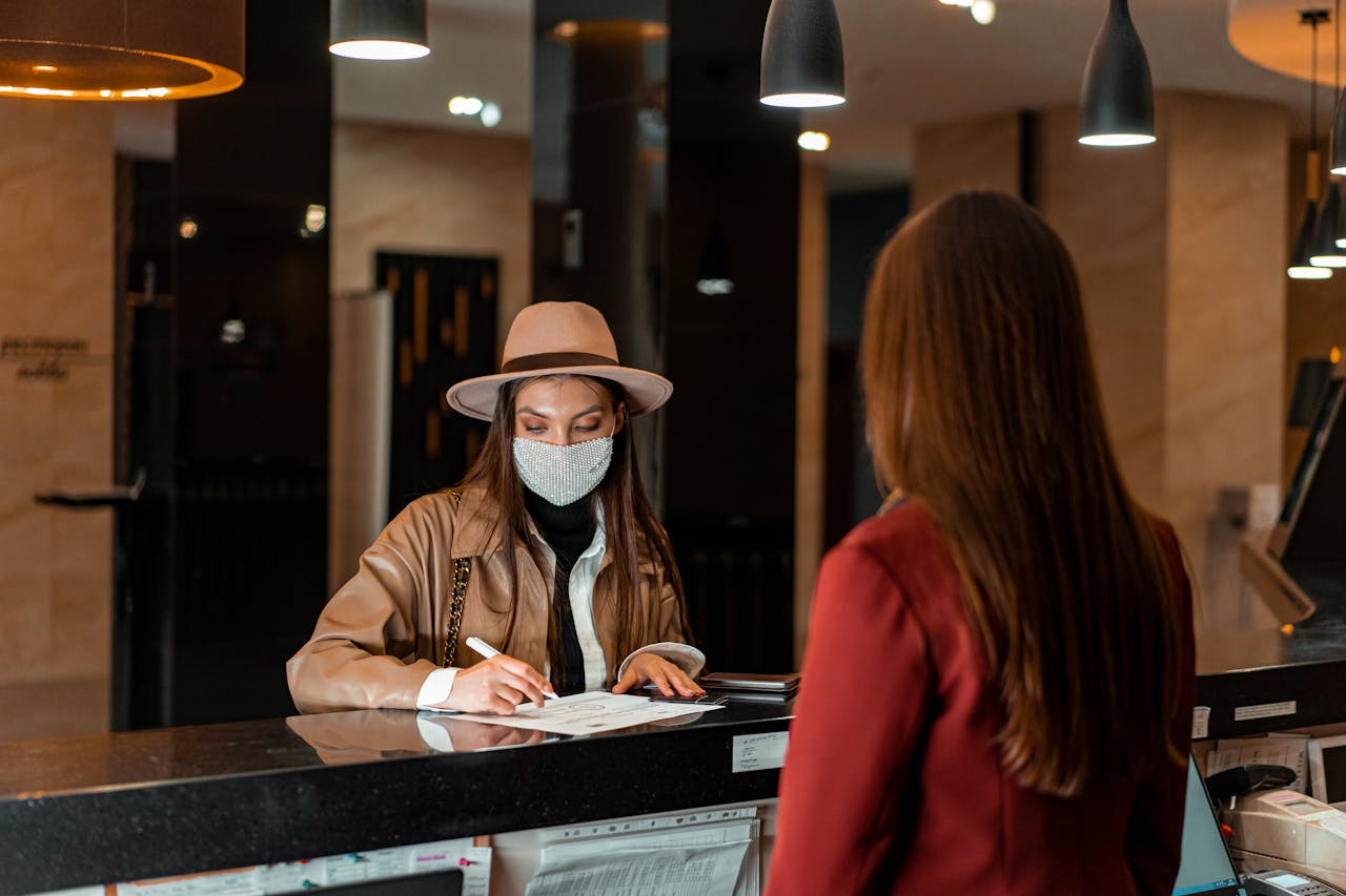 our-services-01 A woman in a hat and face mask checks in at a hotel reception, interacting with a receptionist.
